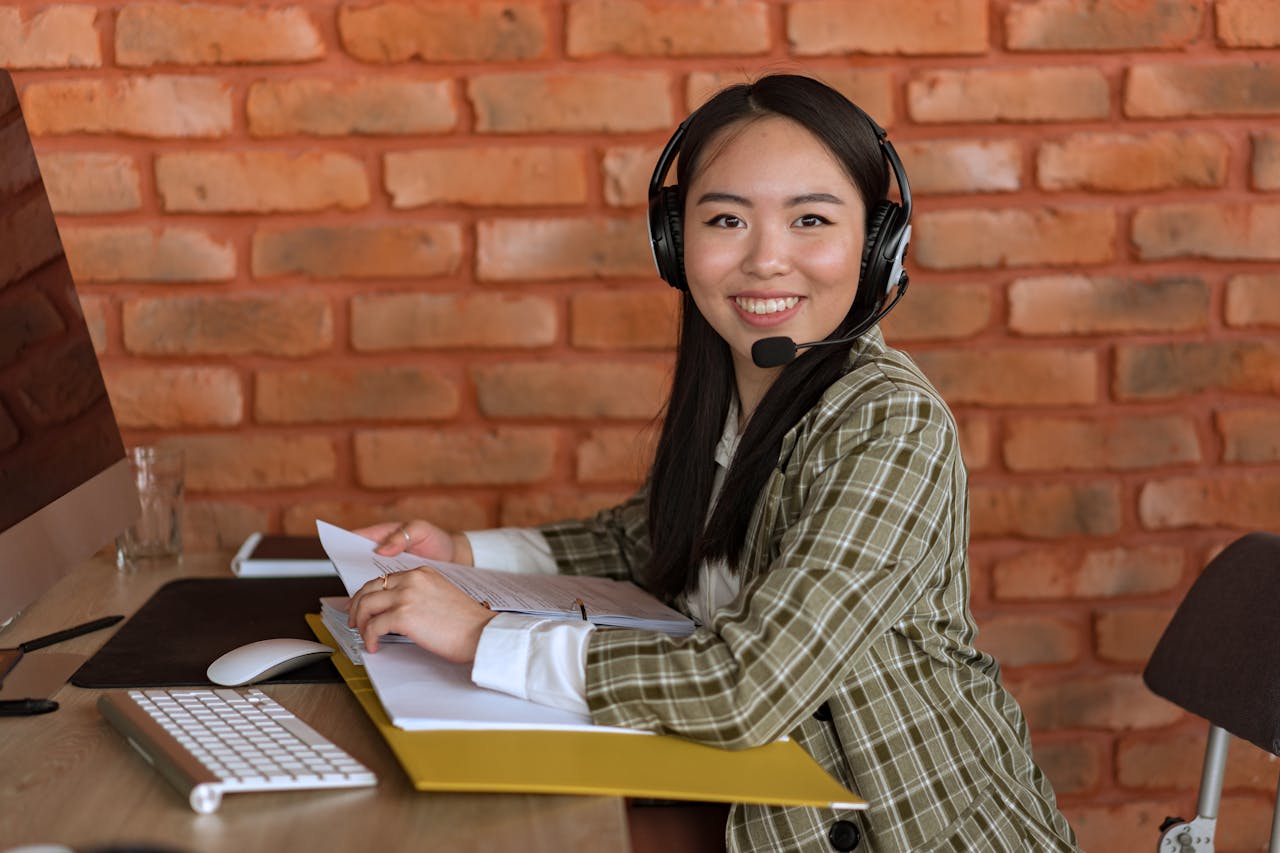 Asian woman working as a call center agent, smiling at desk with documents and headset.